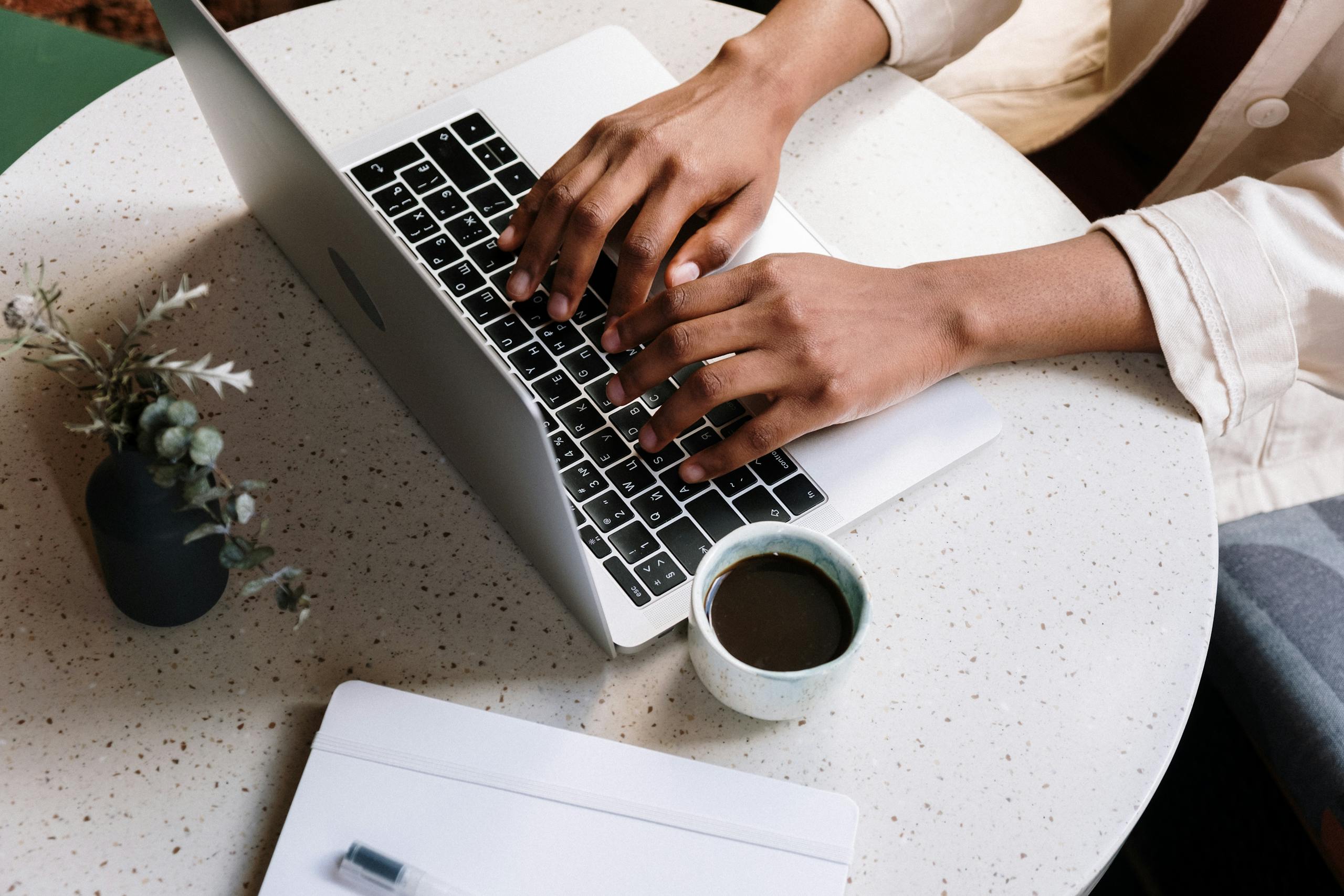 A person typing on a laptop at a café table with coffee and a notebook.